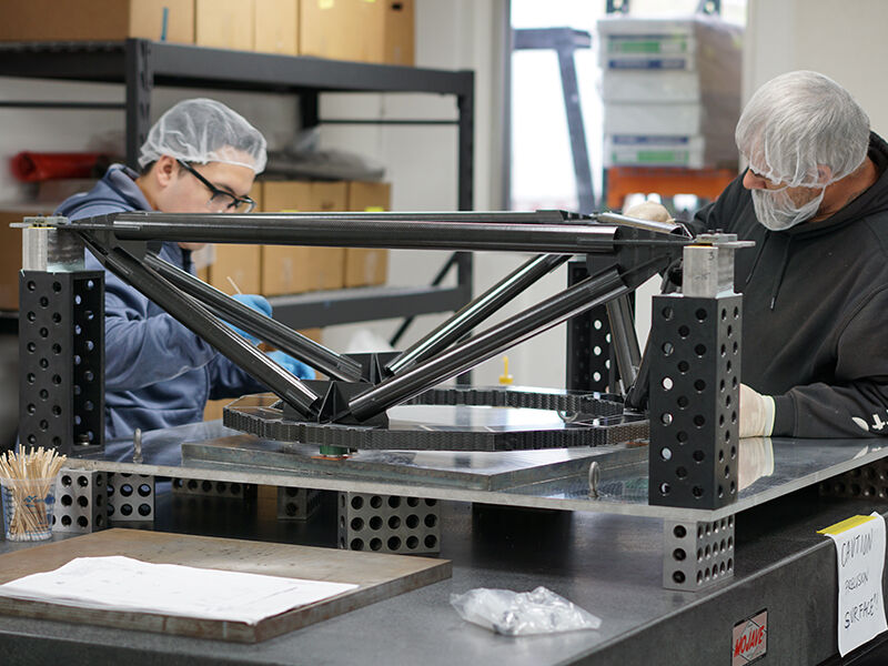 Image shows a manufacturing environment with two technicians working on the TMT metrology fixture. The structure is placed on a metal platform that is sitting on a granite table and held up by three positioning towers. It is comprised of carbon fiber tubes attached at multiple points by dark metallic fittings.